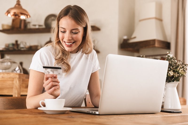 Retrato de una hermosa mujer rubia de 20 años con camiseta casual usando el ordenador portátil y tarjeta de crédito para las compras en línea mientras está sentado en un acogedor café de interior